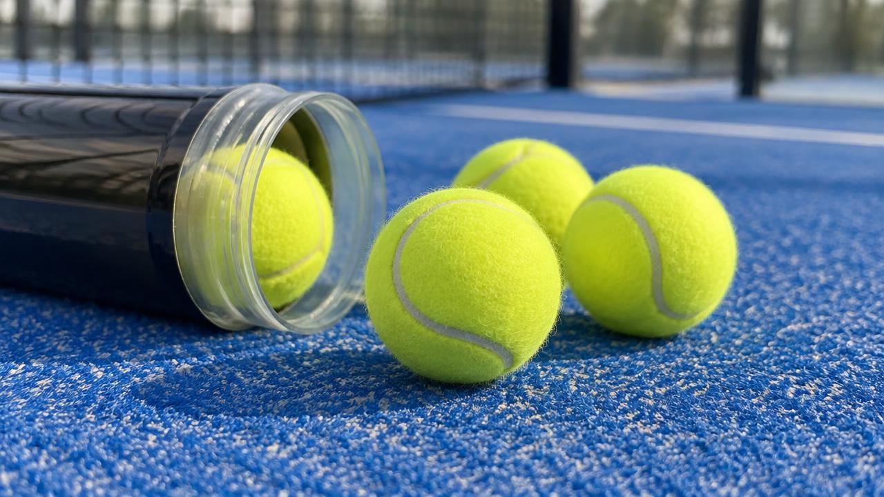 Drei Padel-Bälle neben einer geöffneten Balldose auf blauem Kunstrasen